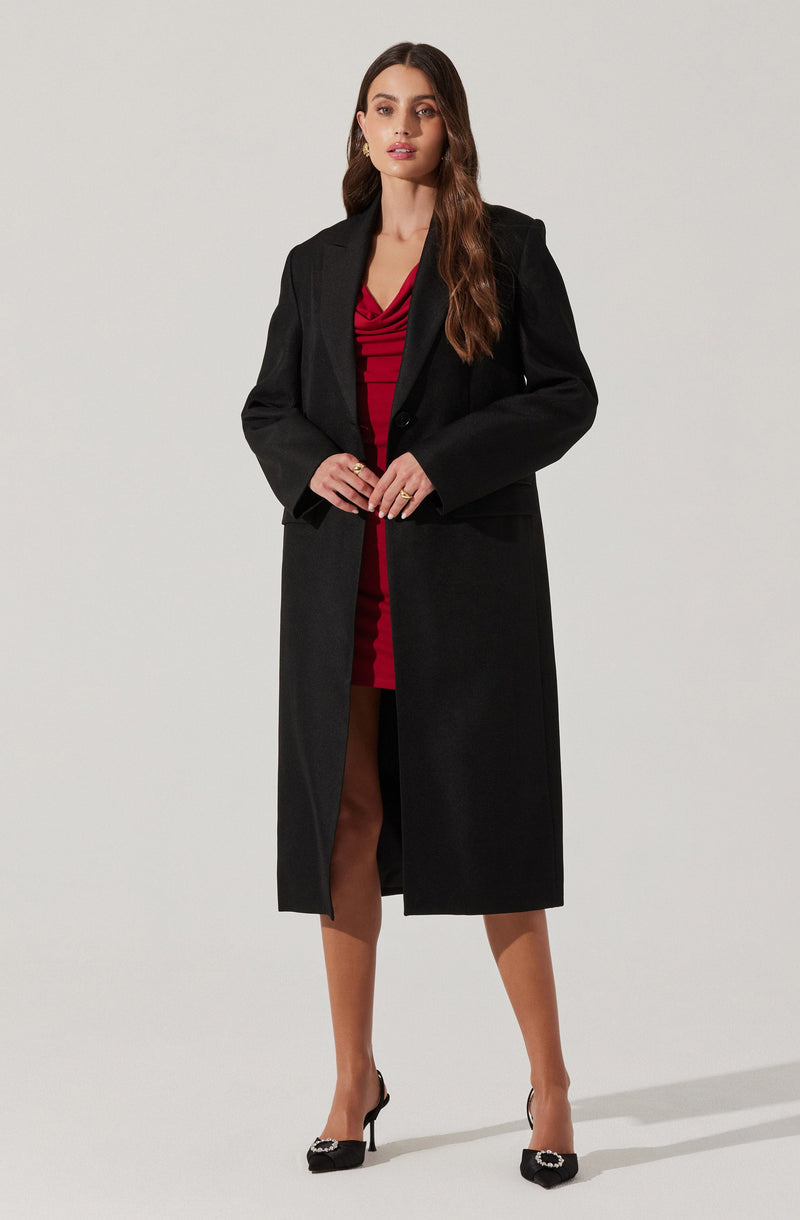 A woman is wearing a long black coat over a red dress with a gathered or ruched bodice. She is also wearing black high heels with decorative embellishments on the toes.