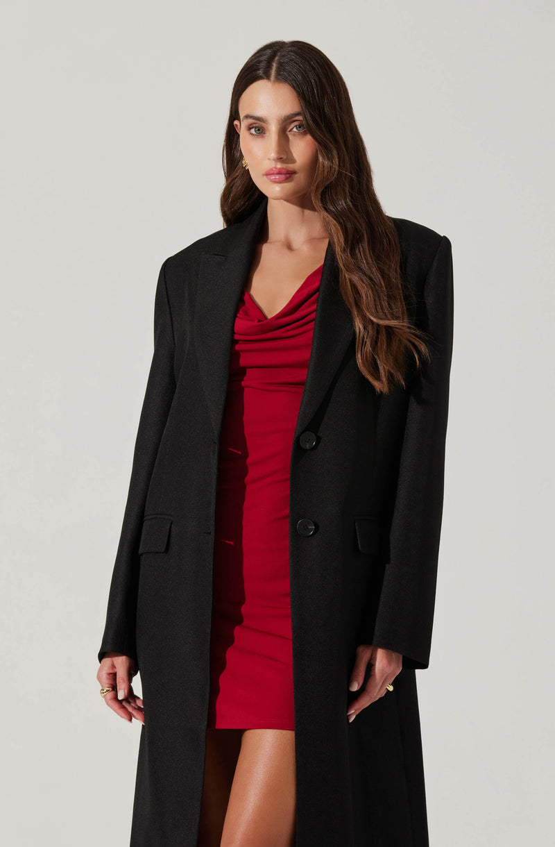 A woman is wearing a fitted, knee-length red dress with a draped neckline. She has on a long black blazer with a notched lapel, two buttons, and flap pockets.