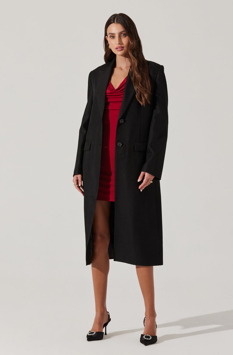 A woman is wearing a long black coat over a red dress with a draped neckline and a short hemline. She is also wearing black high-heeled shoes with embellished details.