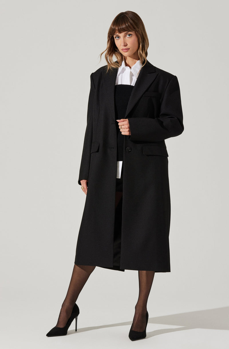 A woman is wearing a long, black blazer-style coat over a white collared shirt and a black sweater. She has black tights and black high-heeled shoes.
