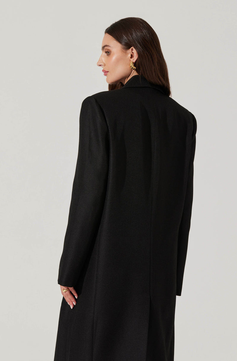 A woman with shoulder-length brown hair is modeling a long black coat. The coat features a structured, tailored fit with a collar and long sleeves, and appears to be made of a textured fabric. She is wearing large, gold hoop earrings. The background is plain and light-colored, emphasizing the coat.
