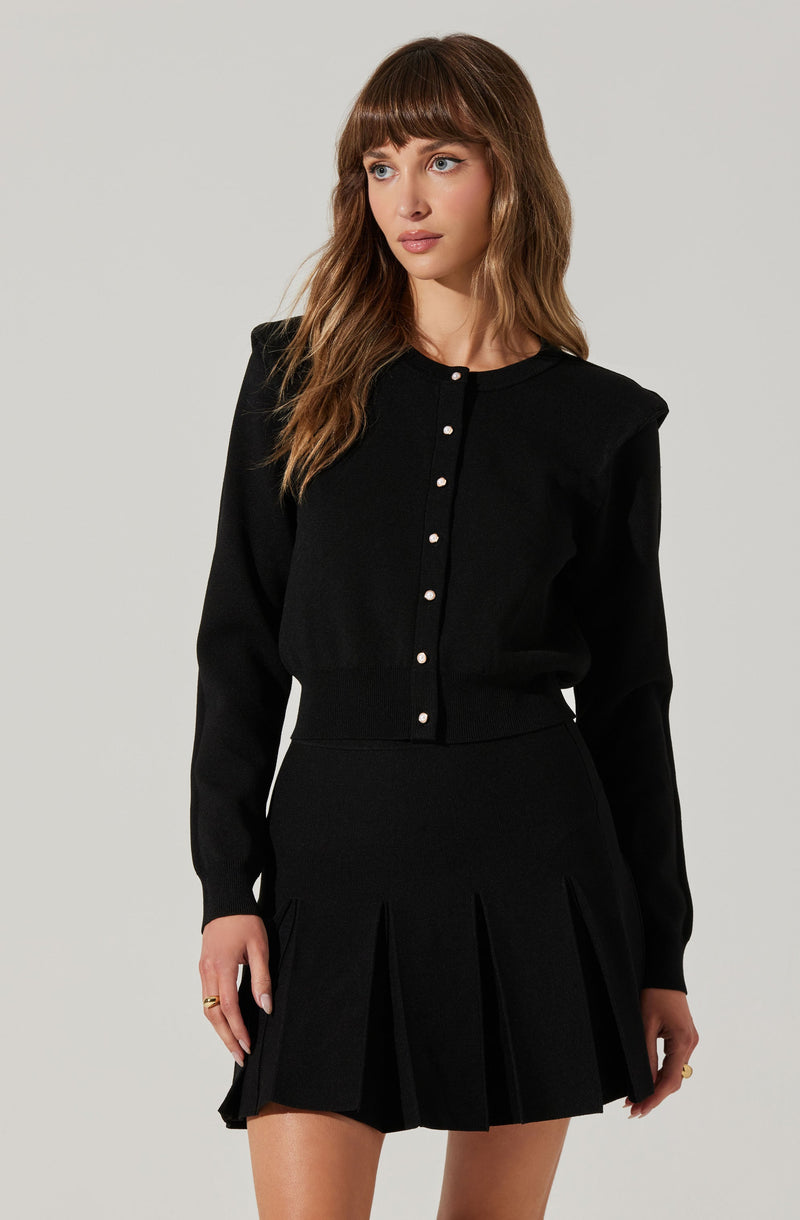 A woman is wearing a black long-sleeved button-up top with pearl-like buttons down the front. The top has slight puff shoulders and a relaxed fit. She pairs it with a high-waisted black pleated skirt that has wide pleats, creating a structured silhouette.