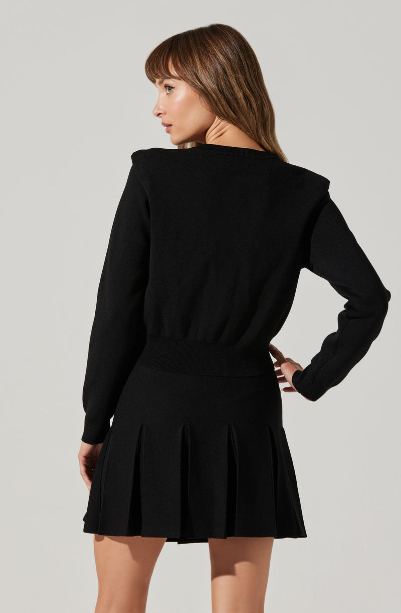 A woman is wearing a black long-sleeve top with a fitted waistband and a black pleated skirt with wide pleats that fall to mid-thigh.