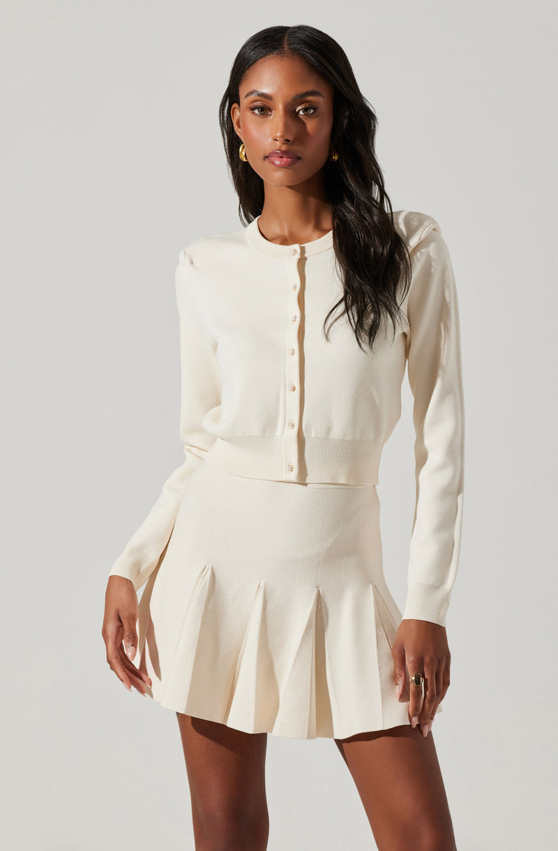 A woman is wearing a cream-colored, long-sleeved, cropped cardigan with small buttons down the front. She is also wearing a matching high-waisted, pleated mini skirt in the same cream color.