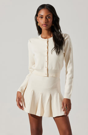A woman is wearing a cream-colored, long-sleeved, cropped cardigan with small buttons down the front. She is also wearing a matching high-waisted, pleated mini skirt in the same cream color.