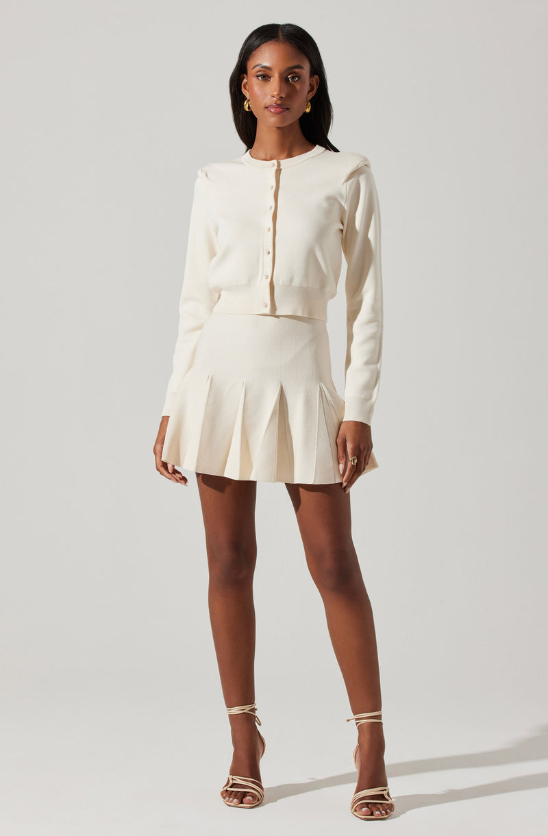 The woman is wearing a cream-colored, long-sleeved cardigan with a round neckline and button-down front. She is also wearing a matching cream-colored pleated skirt with wide pleats that fall above the knee. She has on beige strappy high-heeled sandals and gold earrings.