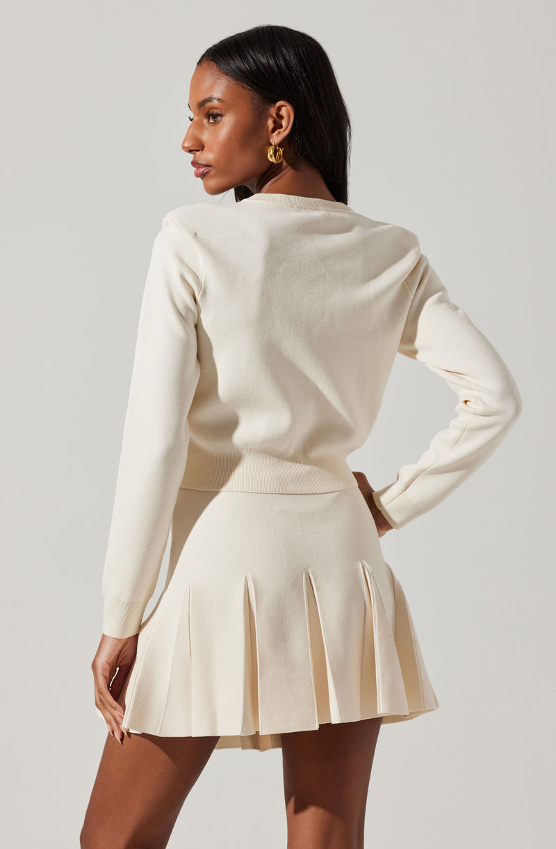 A woman is wearing an all-cream outfit consisting of a long-sleeved, crew neck sweater and a pleated mini skirt. The sweater has a smooth, fitted style, and the skirt features wide, structured pleats. She accessorizes with large gold hoop earrings.