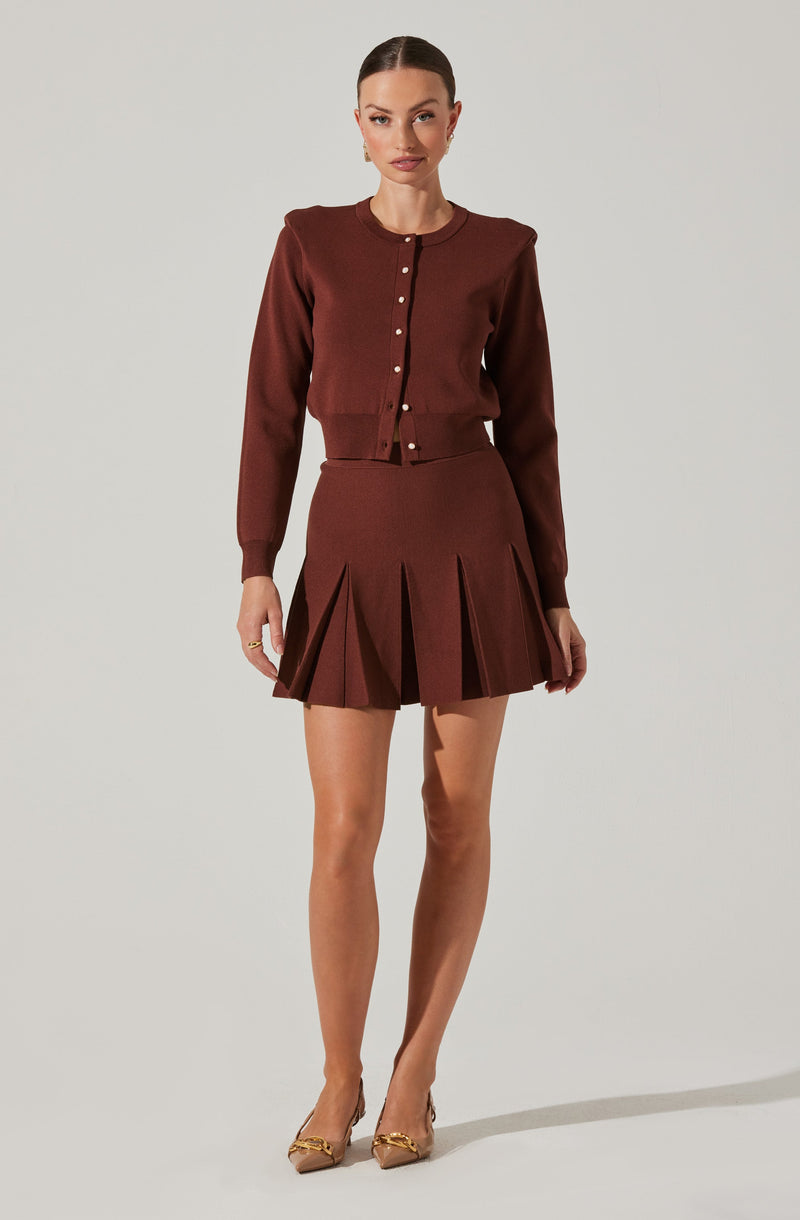 A woman is wearing a matching set consisting of a long-sleeved, button-up cardigan with a ribbed hem and a pleated mini skirt, both in a rich brown color. She is also wearing pointed-toe heels with gold embellishments.