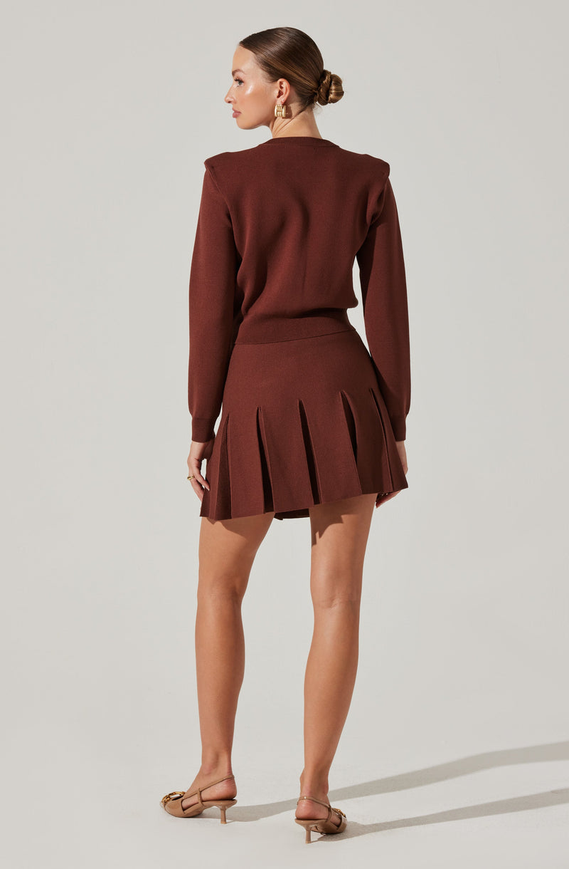 A woman is wearing a matching set of a long-sleeve, brown top and a pleated skirt in the same color. The top appears to be slightly oversized with a round neckline, and the skirt has wide, evenly spaced pleats. She is also wearing beige, open-toed high-heeled shoes with ankle straps.