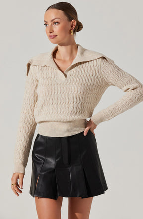 A woman is wearing a cream-colored, cable-knit sweater with a wide collar and a partial zip at the neckline. The sweater has a textured pattern and is slightly loose-fitting. She pairs it with a black, leather mini skirt that features pleats and front slash pockets.