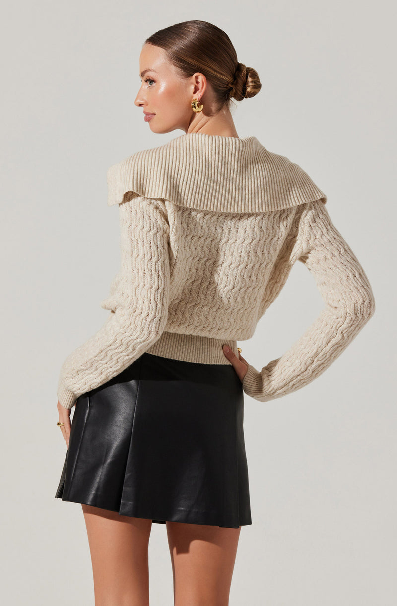 A woman is wearing a cream-colored, chunky knit sweater with a large, ribbed collar that drapes over her shoulders. She is also wearing a high-waisted black leather mini skirt.