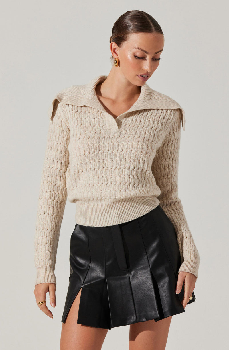 A woman is wearing a cream-colored, cable-knit sweater with a wide collar and shoulder epaulets. She is also dressed in a black, high-waisted leather skirt with front pleats and side slits.