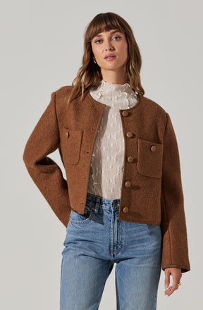 A woman is wearing a brown, cropped, textured jacket with large front pockets and button closures, paired with a white, high-neck blouse with ruffled detailing. She is also wearing high-waisted, light-wash denim jeans.