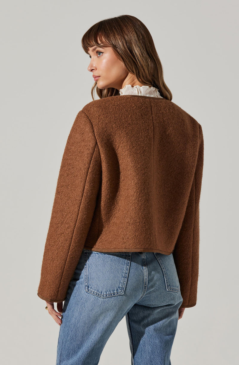 A woman is wearing a brown, textured, long-sleeved, cropped jacket that appears to be made of a wool or similar warm fabric. The jacket has a simple, clean design without visible buttons or embellishments. She pairs it with high-waisted blue jeans. Underneath, she wears a white blouse with a ruffled collar.