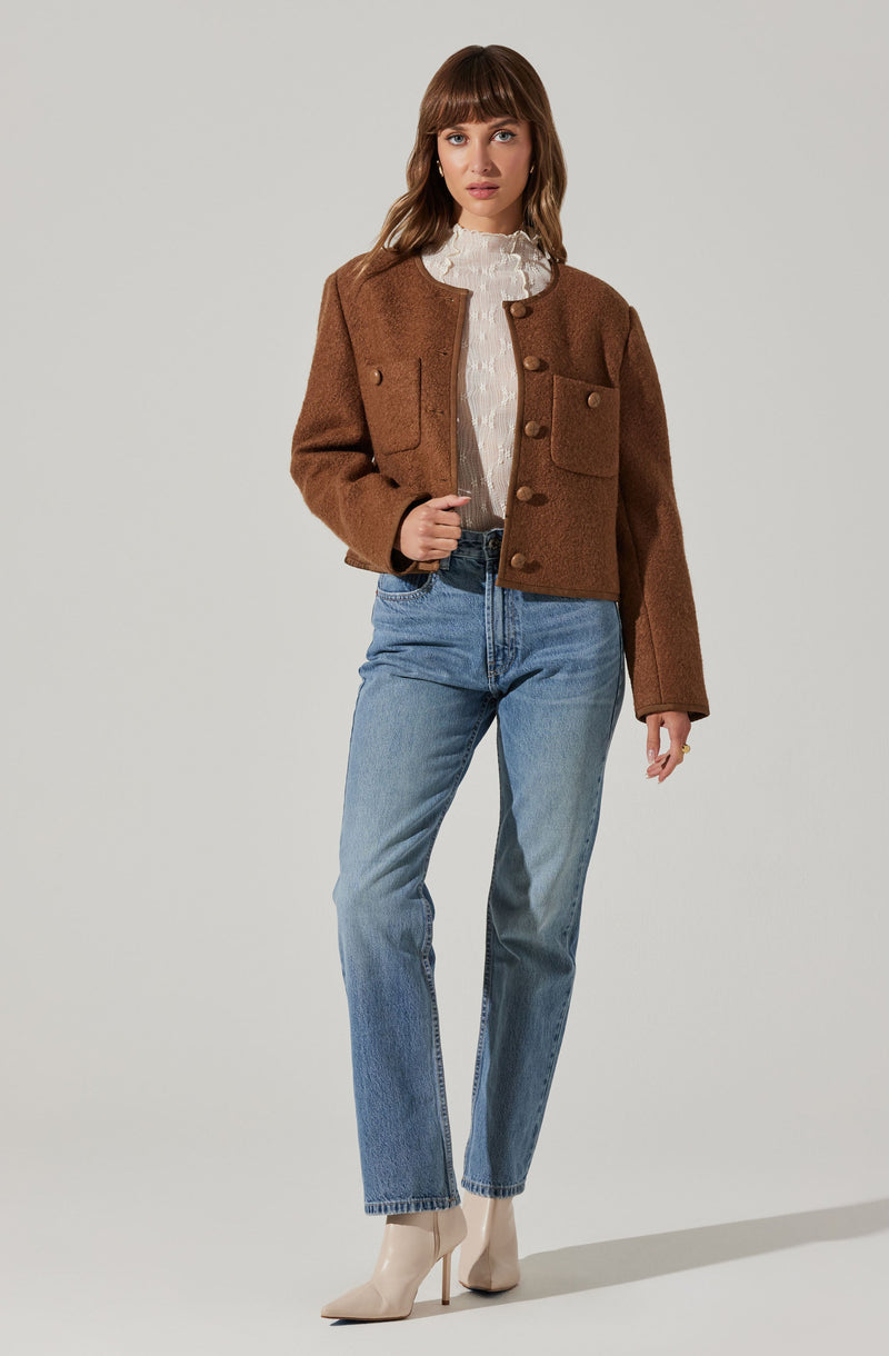 A woman is wearing a cropped brown textured jacket with large front pockets and buttons down the front, paired with a white ruffled high-neck blouse, light blue high-waisted jeans, and beige pointed-toe high-heeled ankle boots.