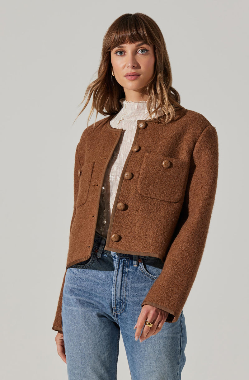 A woman is wearing a brown, textured, cropped jacket with large wooden buttons and two flap pockets on the front. She has on a cream-colored, high-neck blouse underneath and light blue high-waisted jeans.