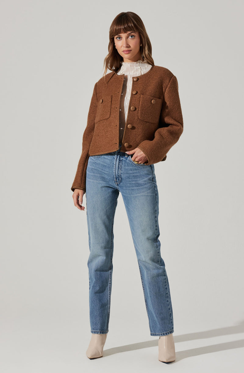The woman is wearing a brown, textured, cropped jacket with large buttons and flap pockets on the chest. Underneath, she has a white, high-neck blouse with ruffle details. She is also wearing light blue, high-waisted jeans with a slightly relaxed fit and rolled-up hems, paired with beige ankle boots.