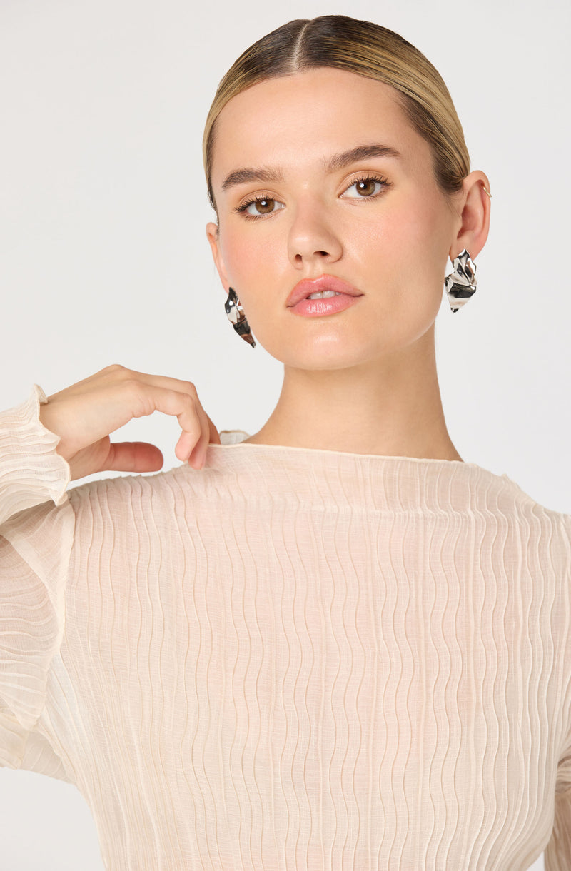 A woman wearing a light beige, long-sleeve, textured blouse with vertical pleats. The blouse has a high, round neckline. She is accessorized with large, bold, twisted silver earrings.