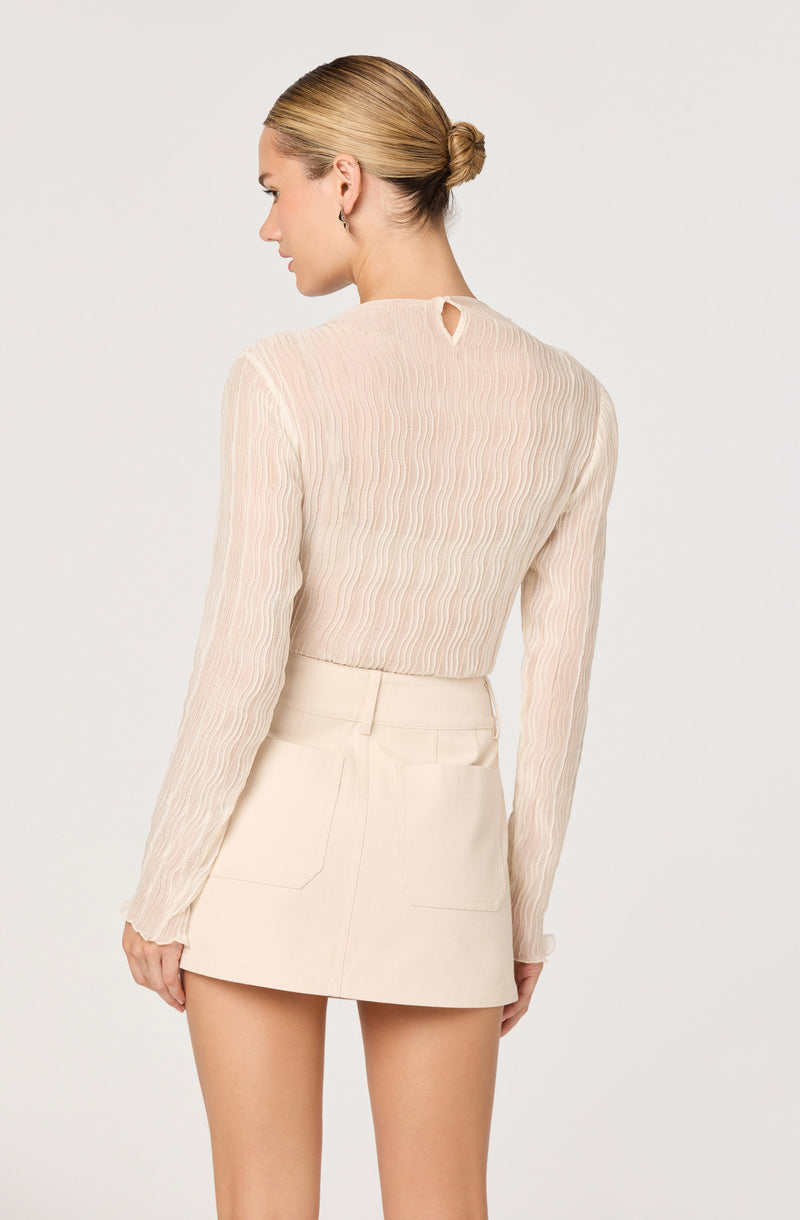A woman is wearing a long-sleeved, sheer, cream-colored blouse with vertical textured stripes and a small keyhole opening at the back neckline. She pairs it with a high-waisted, cream-colored mini skirt featuring two rear patch pockets.