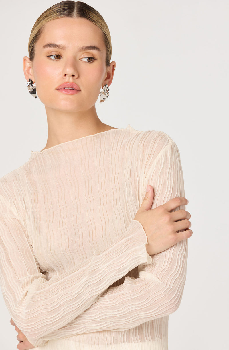 A woman with blonde hair styled straight and pulled back is wearing a light beige, long-sleeved top with a ribbed, textured fabric that features vertical lines. She has on large, bold, black and white statement earrings.
