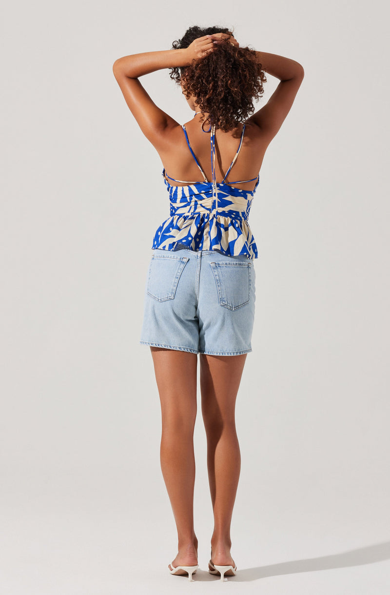 A woman is wearing a blue and white patterned camisole top with spaghetti straps and a ruffled hem. The top features thin straps that crisscross and tie at the back with a visible zipper. She is also wearing light-wash denim shorts with a high waist and two back pockets. She has on white heeled sandals.