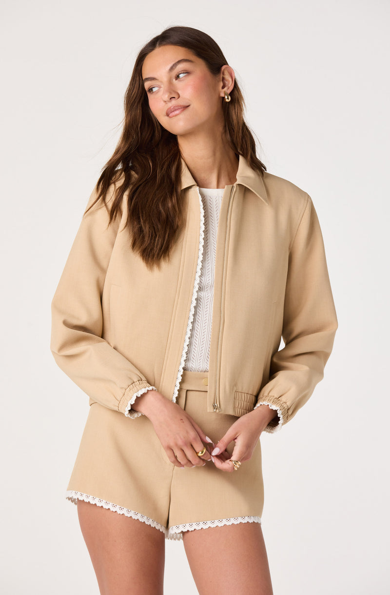 Woman wearing beige jacket and shorts with lace details, and a white top underneath.