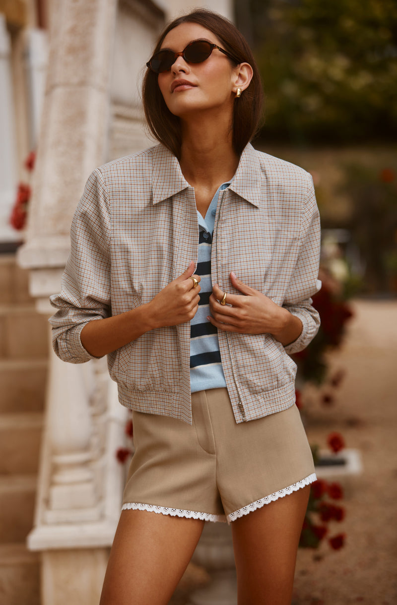 Woman wearing a beige checkered jacket, blue striped shirt, and beige shorts with lace trim.