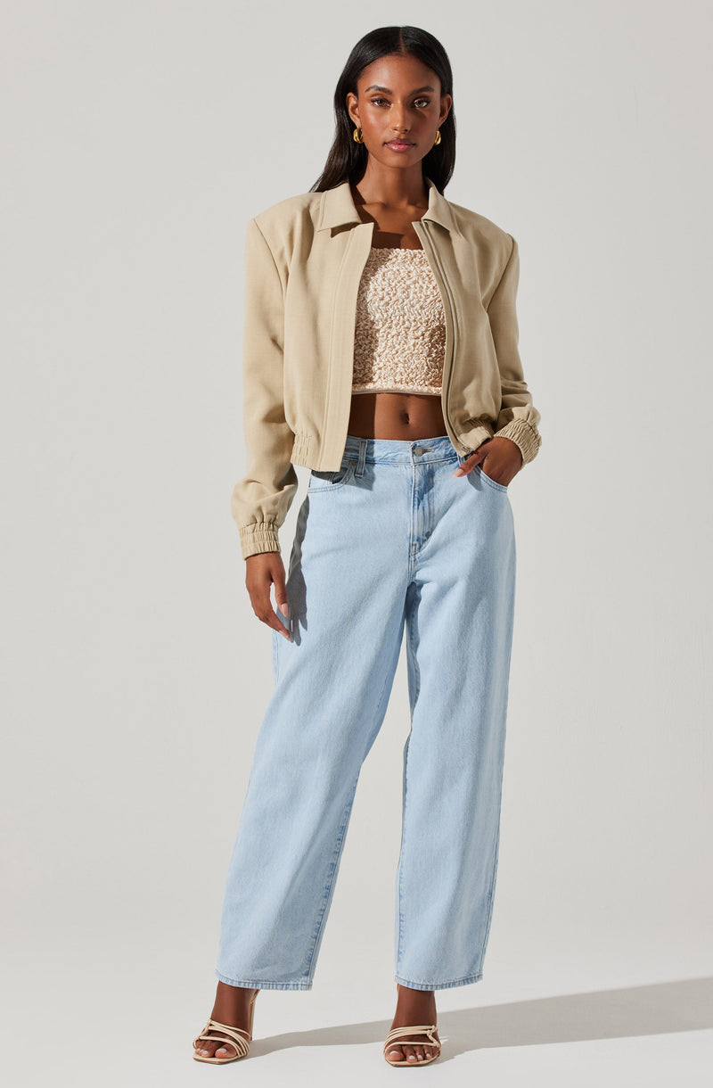 A woman is wearing a beige cropped jacket with ribbed cuffs and a collar, paired with a beige textured crop top. She has on light blue high-waisted, wide-leg jeans and beige strappy high-heeled sandals. She is accessorized with gold earrings.