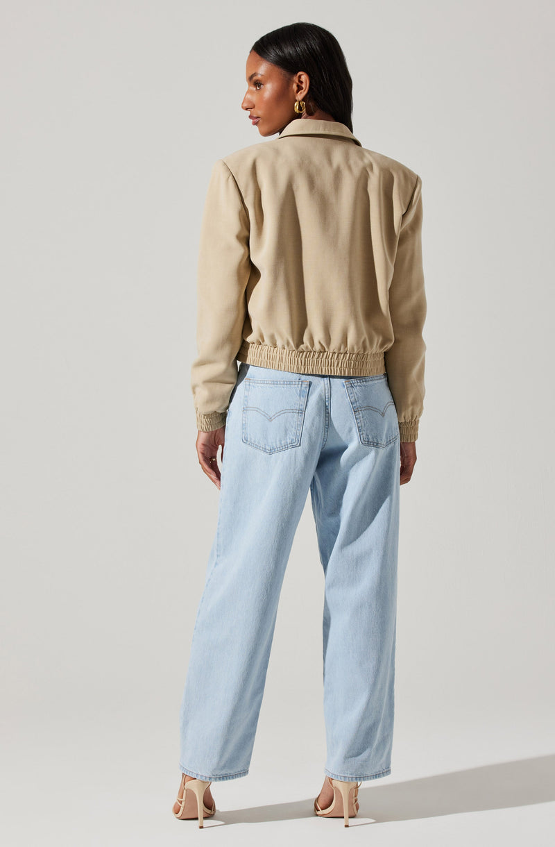 A woman is wearing a beige bomber jacket with a gathered waistband, relaxed long sleeves, and a collar. She is also wearing light blue, high-waisted, wide-leg jeans with visible stitching on the back pockets. She has on beige high-heeled sandals and gold hoop earrings.