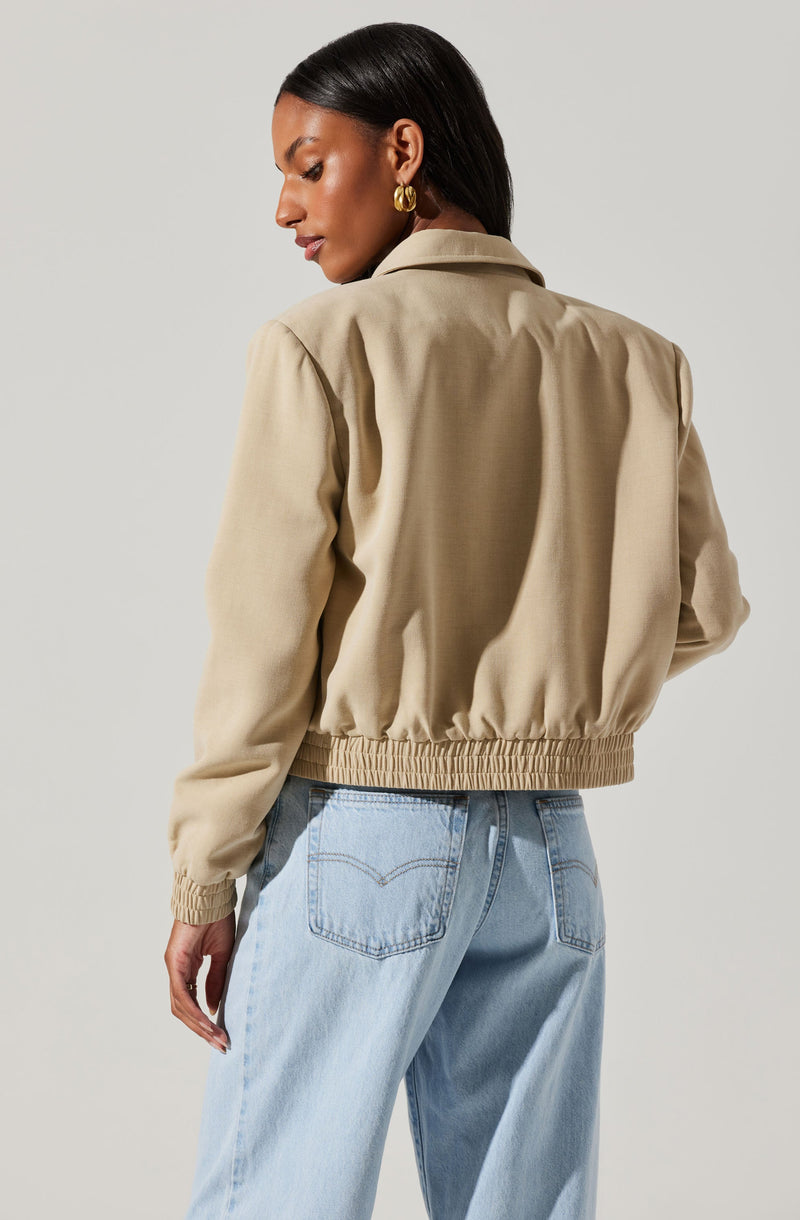 A woman is wearing a beige bomber jacket with a loose fit, gathered at the waist with an elastic band. The jacket has a shirt collar, long sleeves, and appears to be made of a soft, lightweight fabric. She is also wearing light blue high-waisted jeans with a relaxed fit and visible back pockets. The woman has dark, straight hair and is accessorized with large gold hoop earrings.