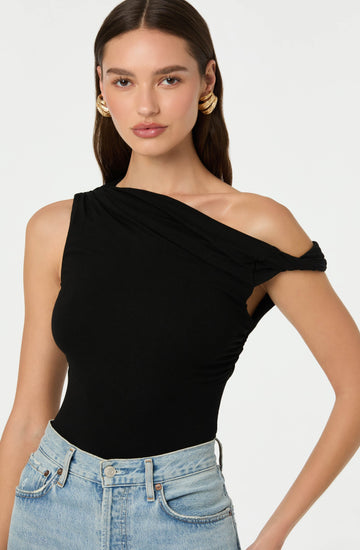 A woman is wearing a black one-shoulder top with ruching on the shoulder strap, paired with light blue high-waisted jeans. She has long, dark brown hair and is accessorized with large, gold hoop earrings.