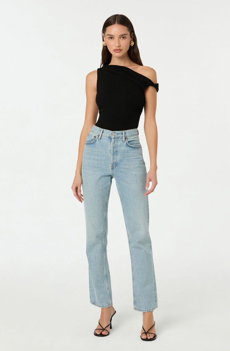 A woman is wearing a black, one-shoulder top with a unique folded and draped design on the neckline and shoulder. She pairs the top with high-waisted, light-wash straight-leg jeans that have a relaxed fit. She completes the look with black, strappy high-heeled sandals.
