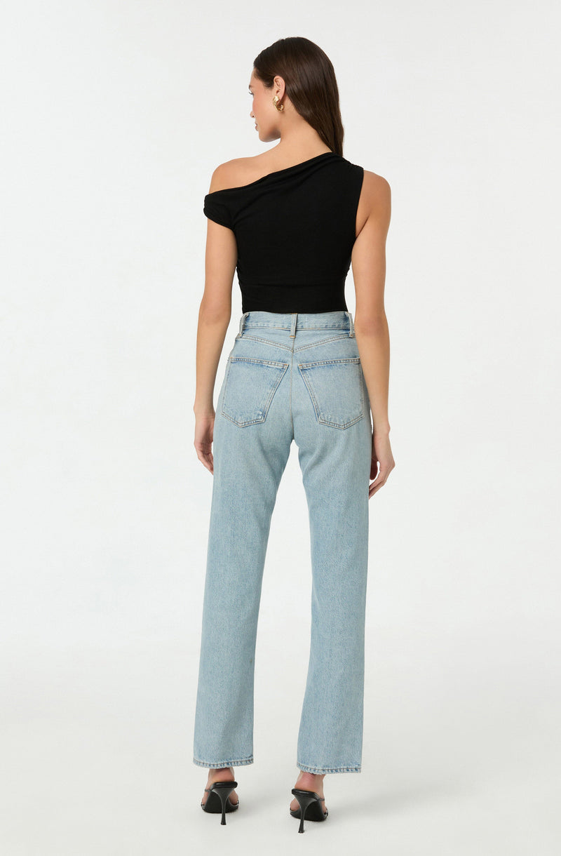 A woman is wearing a black, one-shoulder, short-sleeved top paired with high-waisted, light-wash, straight-leg jeans. She is also wearing black high-heeled shoes with ankle straps.