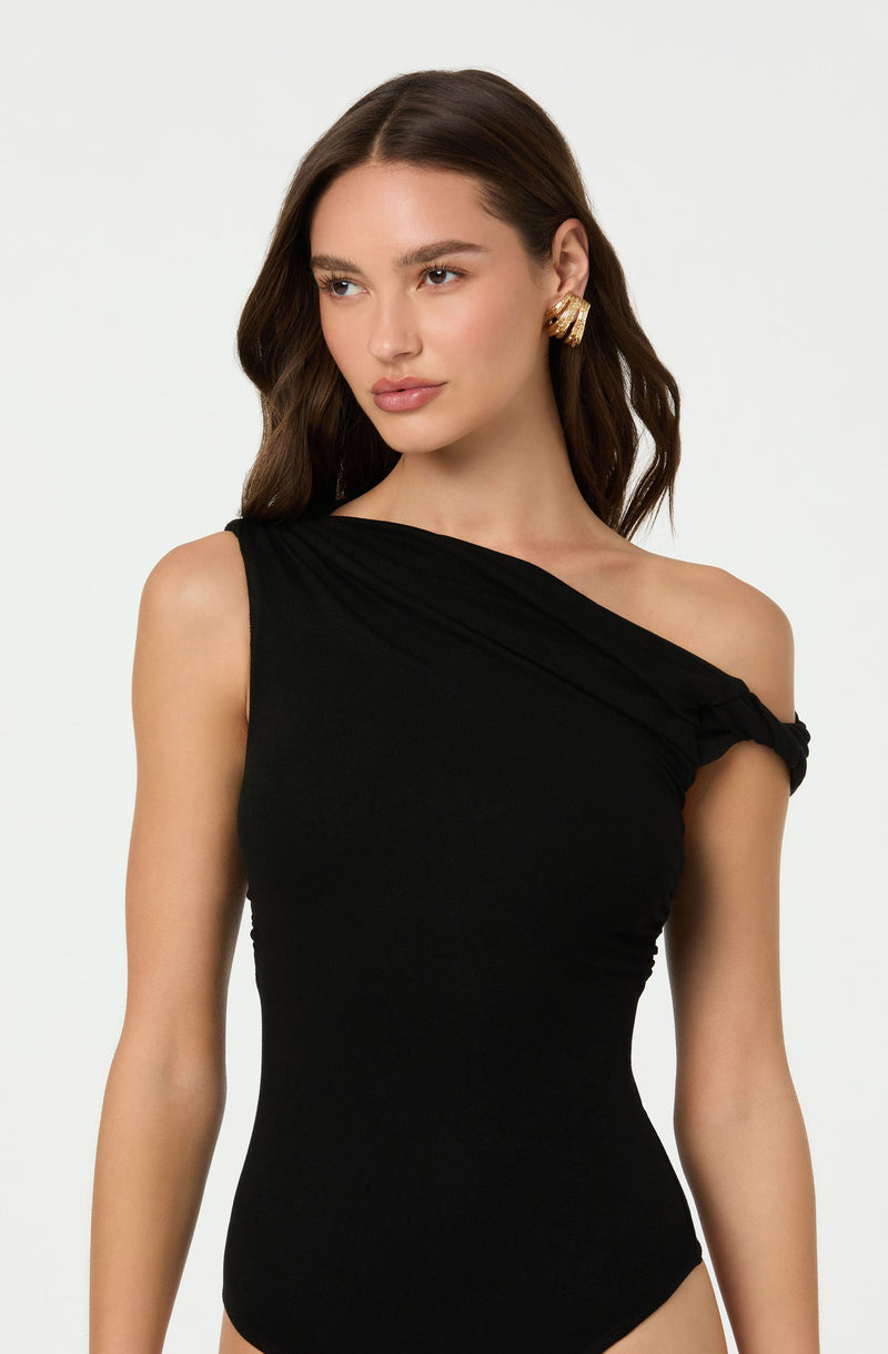 A woman is wearing a black, one-shoulder bodysuit with a gathered detail on the shoulder strap. She accessorizes with large, gold hoop earrings.