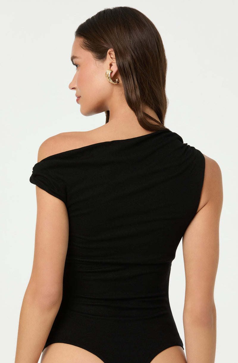 A woman is wearing a black, one-shoulder top with a draped, ruched design. The top exposes her left shoulder and has a fitted silhouette. She is accessorized with a large, gold hoop earring.