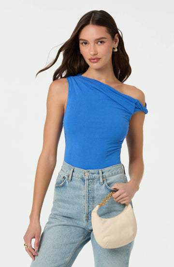 A woman is wearing a bright blue sleeveless top with an asymmetrical design and a tied strap detail on one shoulder. She pairs it with high-waisted, light-wash jeans. She is holding a small, off-white handbag with a gold chain strap.
