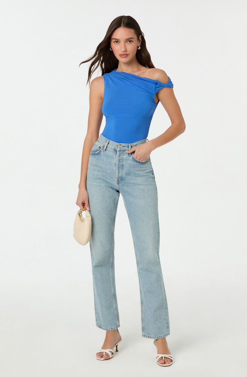 A woman is wearing a bright blue sleeveless top with draped fabric detail on the shoulders, paired with high-waisted, light-wash, straight-leg jeans. She accessorizes with small gold earrings, a small off-white handbag, and white high-heeled sandals.