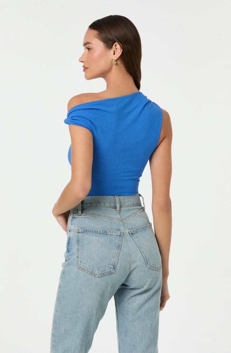 A woman is wearing a blue, one-shoulder top with a draped sleeve on one side, paired with high-waisted, light-wash jeans that have back pockets.