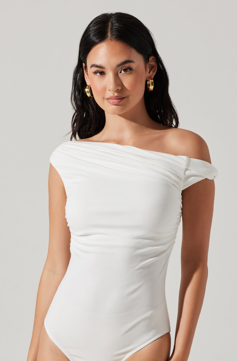 A woman is wearing an off-the-shoulder white gown made of smooth, slightly stretchy fabric. The gown features a gathered detail across the neckline and shoulder, creating a draped effect, with the fabric ruched along the sides for a fitted silhouette. She is accessorized with large gold hoop earrings.