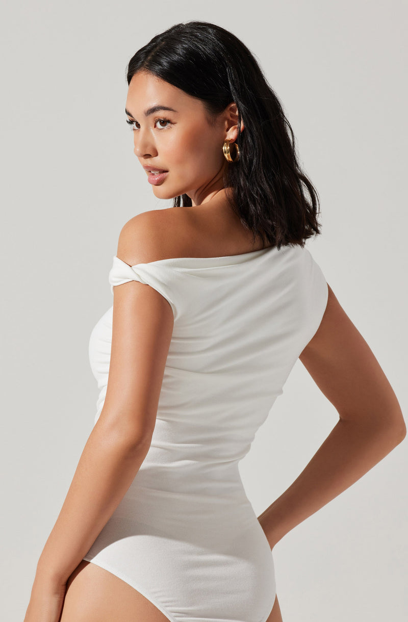A woman is wearing a white, off-the-shoulder dress made of soft, smooth fabric. The dress is form-fitting and has a slight sheen, with a draped detail over the shoulder. She accessorizes with large, gold hoop earrings.