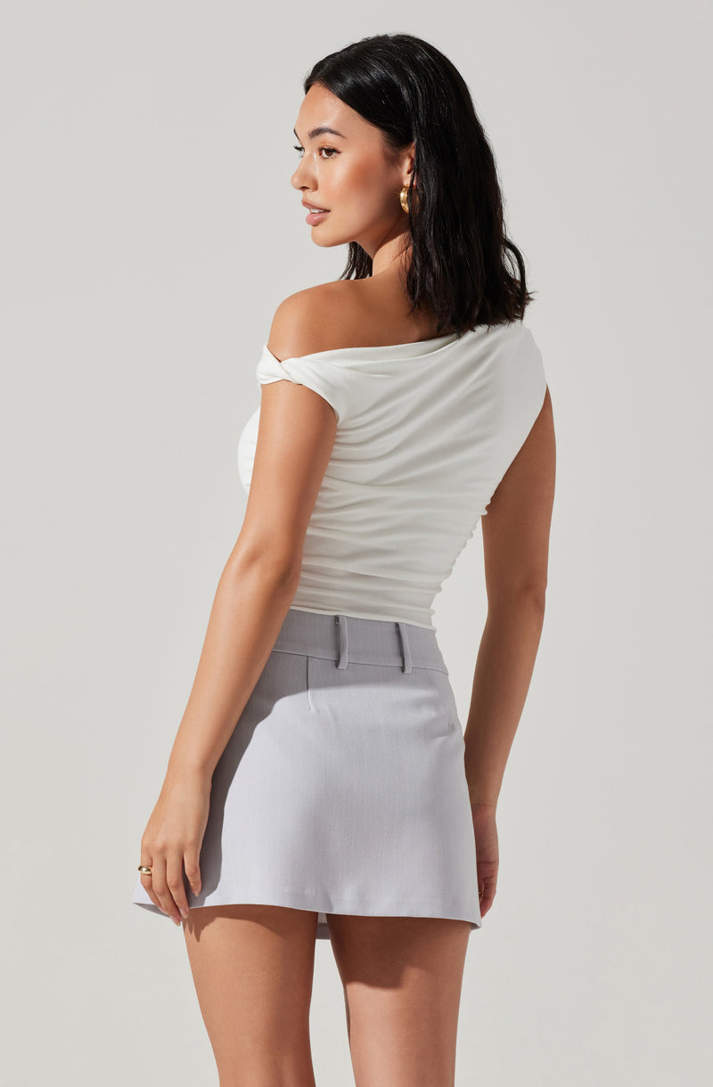 A woman is wearing an off-the-shoulder, short-sleeved, cream-colored top with ruching details, paired with a light gray, high-waisted mini skirt.