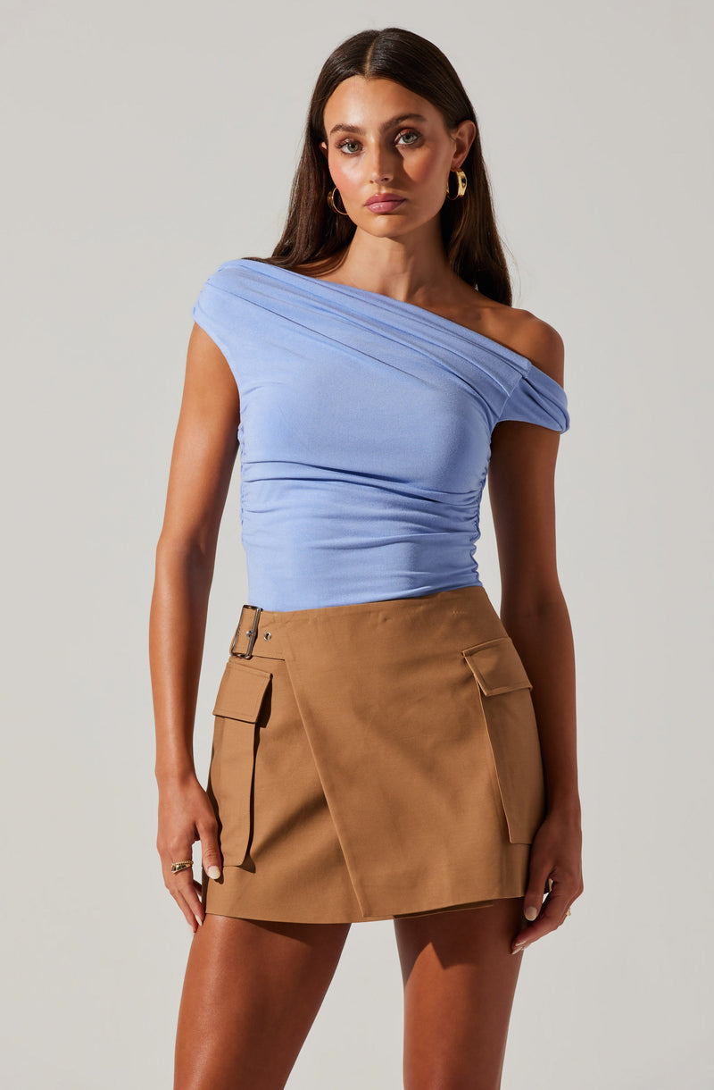 A woman is wearing a light blue, one-shoulder top with gathered fabric details, paired with a high-waisted, tan mini skirt featuring large front pockets with flaps and a belt loop.