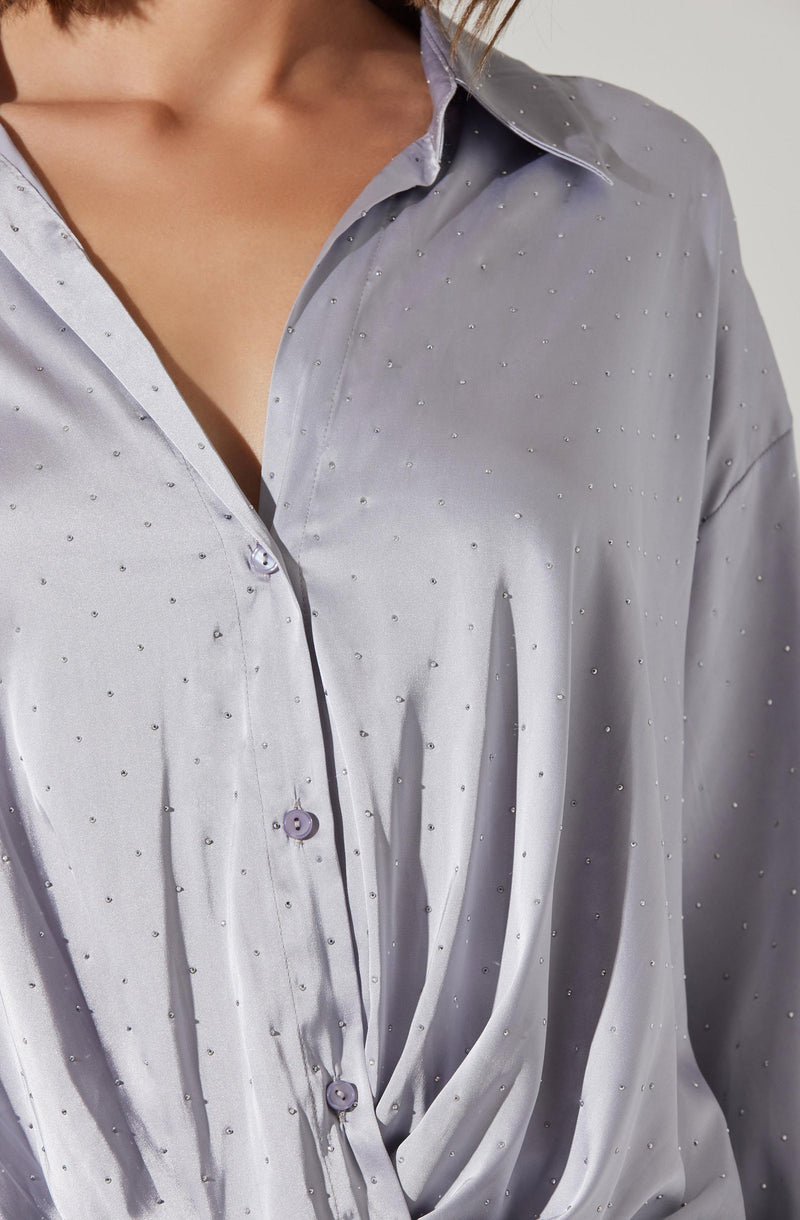 A close-up of a light gray satin button-up shirt featuring small, evenly spaced, shimmering embellishments. The shirt has a classic collar and is worn slightly open at the top. The fabric appears smooth and glossy, with soft pleats creating a tailored look.