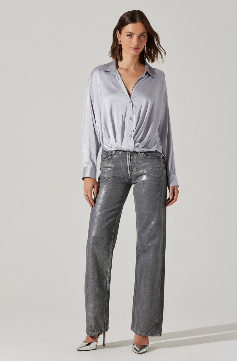 A woman is wearing a light gray satin button-up shirt with a loose fit, long sleeves, and a classic collar. She has tucked the shirt slightly into her high-waisted, metallic silver jeans that have a straight-leg cut. She is also wearing pointed-toe high heels with a metallic finish.