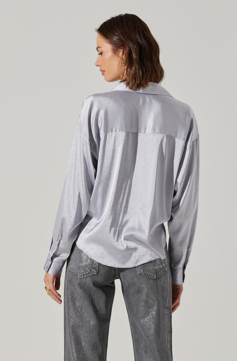 A woman is wearing a light gray satin shirt with a subtle dotted pattern. The shirt has long sleeves, a collar, and a yoke across the back. She is also wearing high-waisted, faded gray jeans.