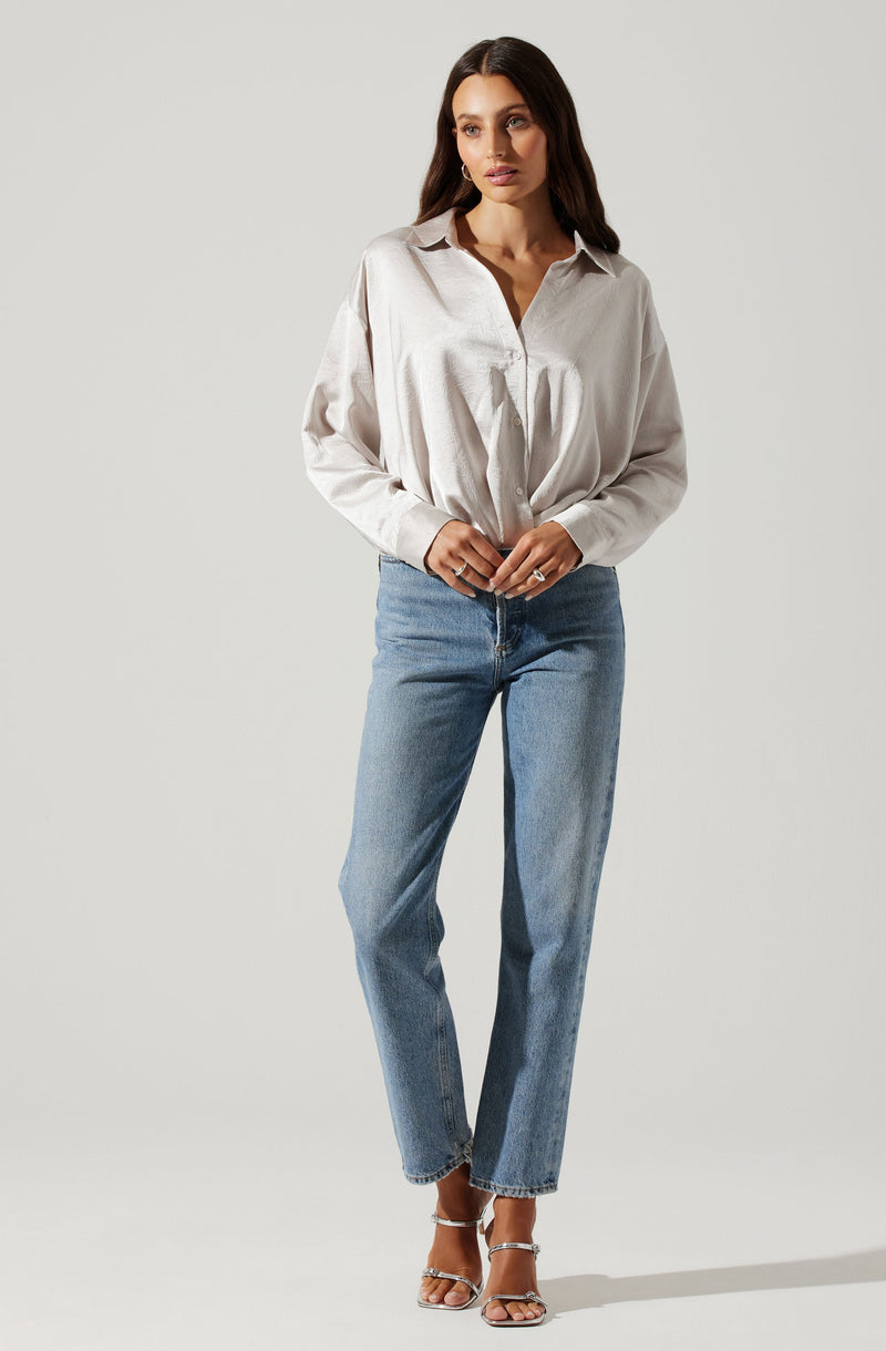 A woman is wearing a loose-fitting, long-sleeved, button-up shirt with a collar made of a silky, shiny fabric in a light cream or off-white color. She has paired it with high-waisted, straight-leg blue jeans and silver strappy high-heeled sandals.