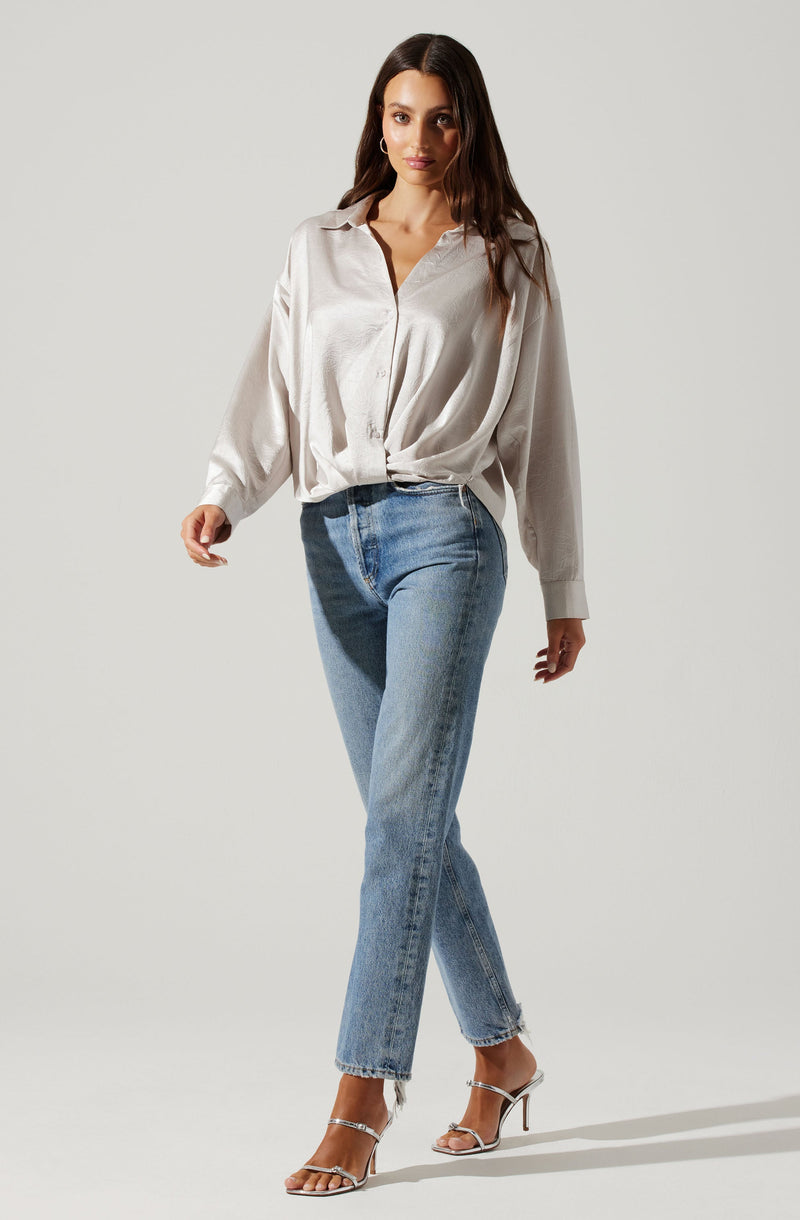 A woman is wearing a loose, long-sleeved, button-up blouse in a champagne or light beige color, tucked slightly at the front. She pairs it with high-waisted, light blue straight-leg jeans and silver high-heeled sandals.