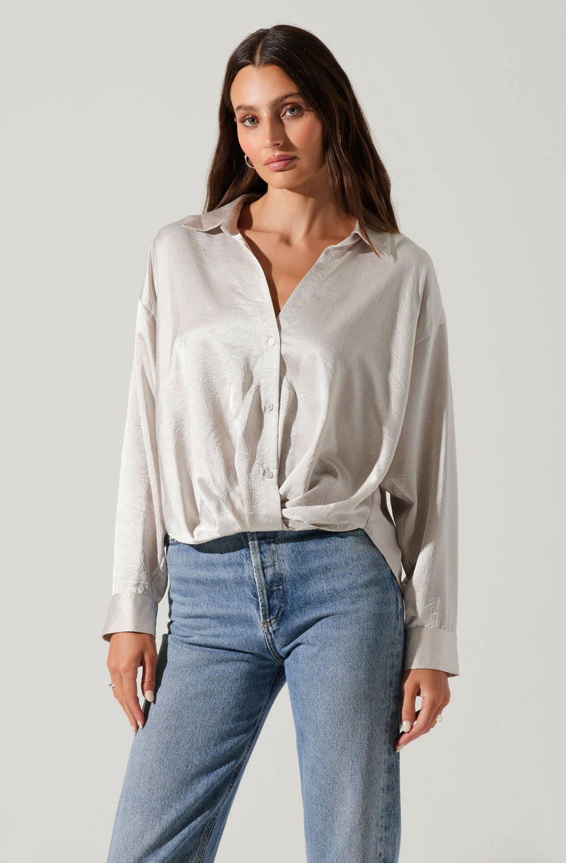 A woman is wearing an oversized, silky white button-up shirt with a collar and long sleeves. The shirt has a shiny texture and is partially tucked into light blue, high-waisted jeans.