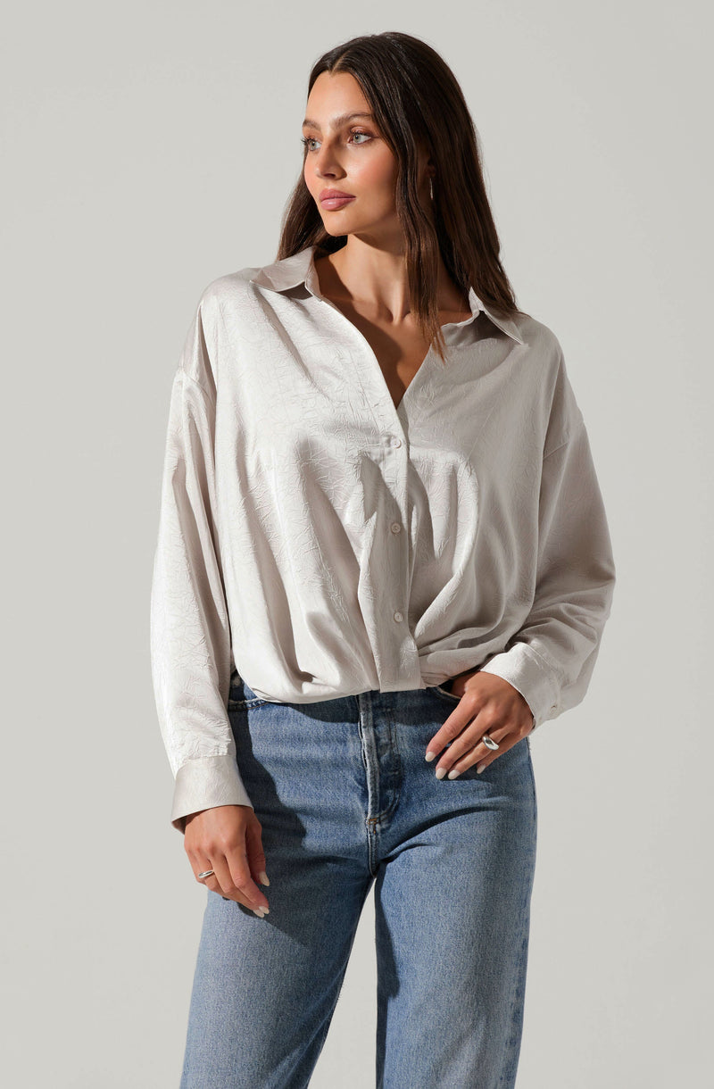 A woman is wearing an oversized, long-sleeved, light-colored button-up shirt with a collar. The shirt appears to be made of a soft, slightly shiny fabric and is loosely tucked into her high-waisted blue jeans.