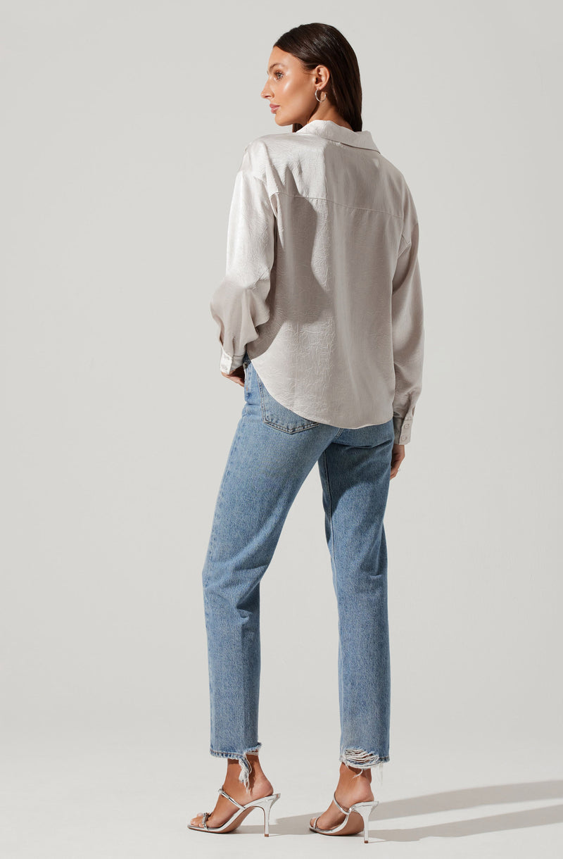 A woman is wearing a loose-fitting, light-colored, long-sleeved button-up shirt with a collar. She pairs it with high-waisted, light blue, slightly distressed jeans that have frayed hems at the ankles. She completes her outfit with strappy, high-heeled sandals.